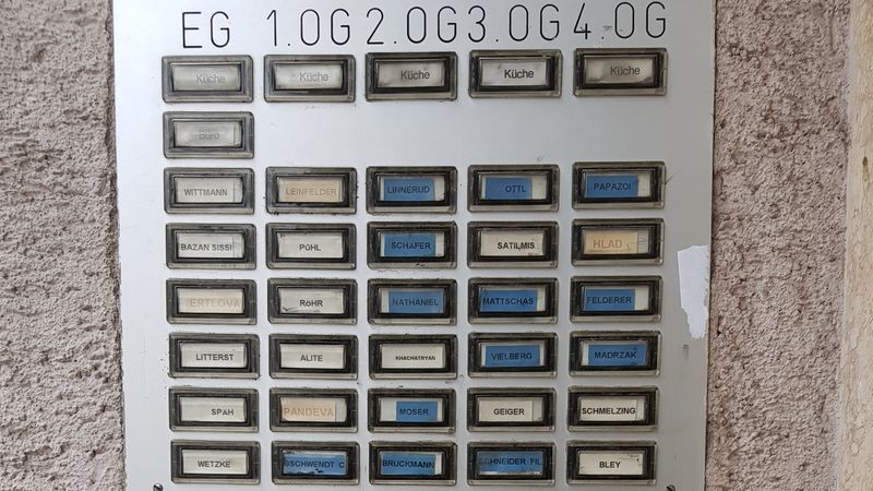 Auf dem Bild ist eine Hausklingelanlage zu sehen, die aus einer Vielzahl von Klingelschildern besteht und an einer Wand außen angebracht ist.