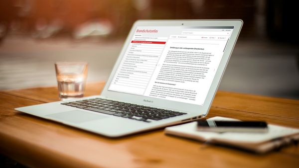 Ein Laptop auf einem Holztisch zeigt ein geöffnetes Textdokument. Daneben steht ein Glas Wasser und ein Notizbuch mit Stift.